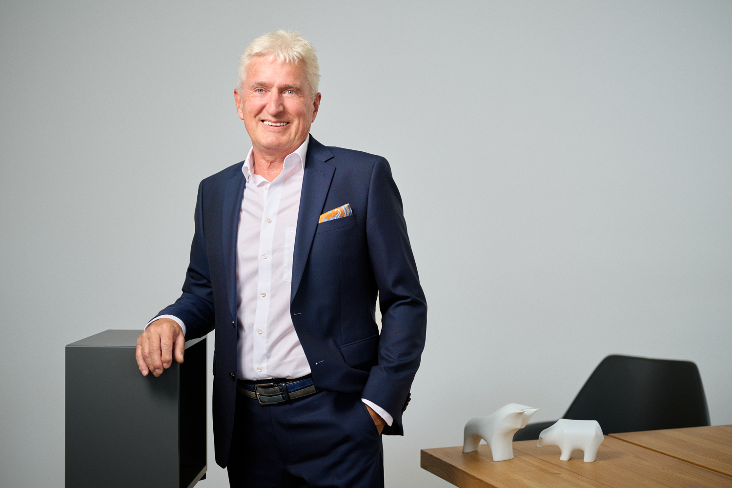 Portrait von Bernhard Roggenhofer: ein älterer Mann mit weißem Haar in dunkelblauem Anzug und weißem Hemd steht lächelnd in einem modernen Büro. Eine Hand ruht auf einem dunklen Möbelstück, im Hintergrund ein Tisch mit weißen Stier- und Bärfiguren.