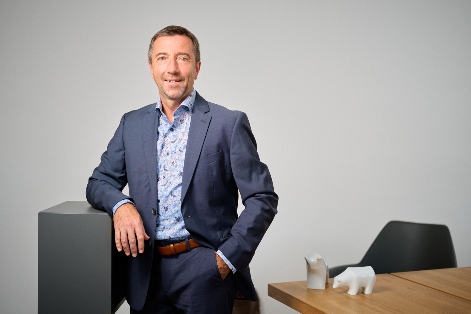 Portrait von Manfred Hübner: ein Mann mittleren Alters mit kurzem braun-grauem Haar, gekleidet in einen dunkelblauen Anzug mit gemustertem Hemd, lehnt entspannt an einem Möbelstück in einem modernen Büro. Im Hintergrund stehen weiße Stier- und Bärfiguren auf einem Holztisch.
